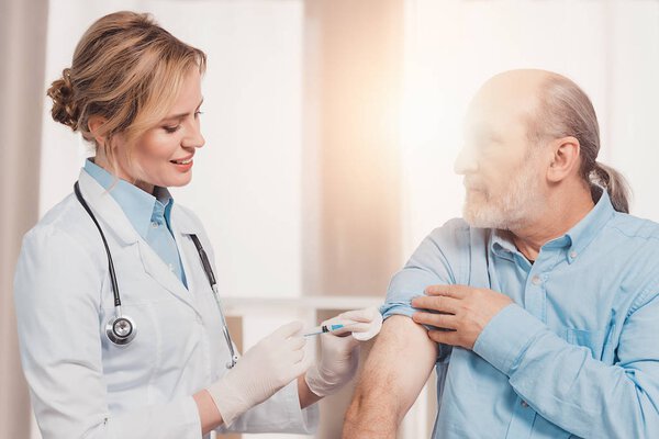 smiling doctor in white coat making injection to senior patient in clinic