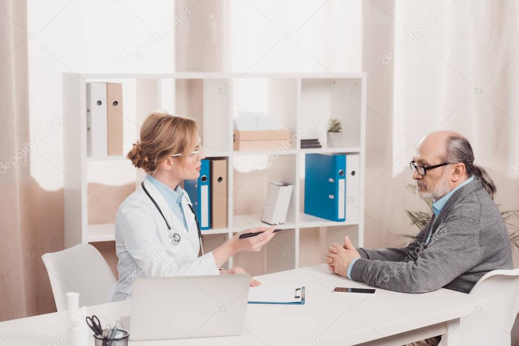 side view of senior patient and doctor having conversation at doctors workplace in clinic