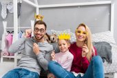 happy young family with masquerade crowns and eyeglasses sitting in kid bedroom