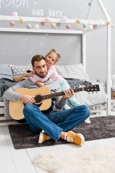 father playing guitar for daughter on floor of kid bedroom while she embracing him