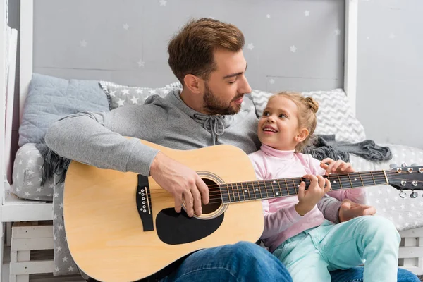 father playing guitar for daughter on floor of kid bedroom