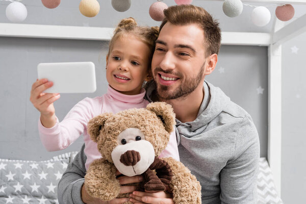 happy young father and daughter with teddy bear taking selfie