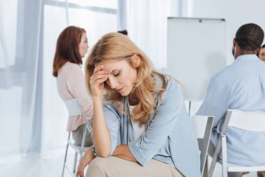 upset mature woman looking down while multiethnic group sitting behind  clipart