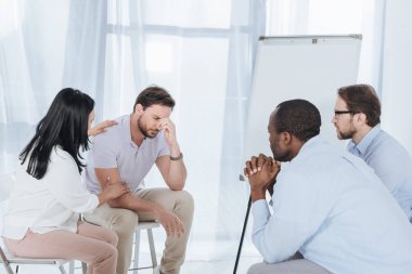 multiethnic middle aged people sitting on chairs and supporting upset man during anonymous group therapy   clipart