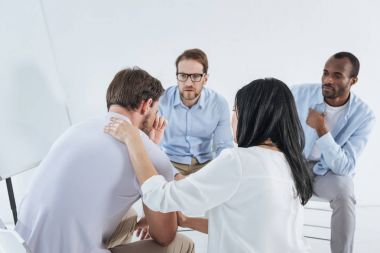 multiethnic mid adult people sitting on chairs and supporting upset man during anonymous group therapy    clipart