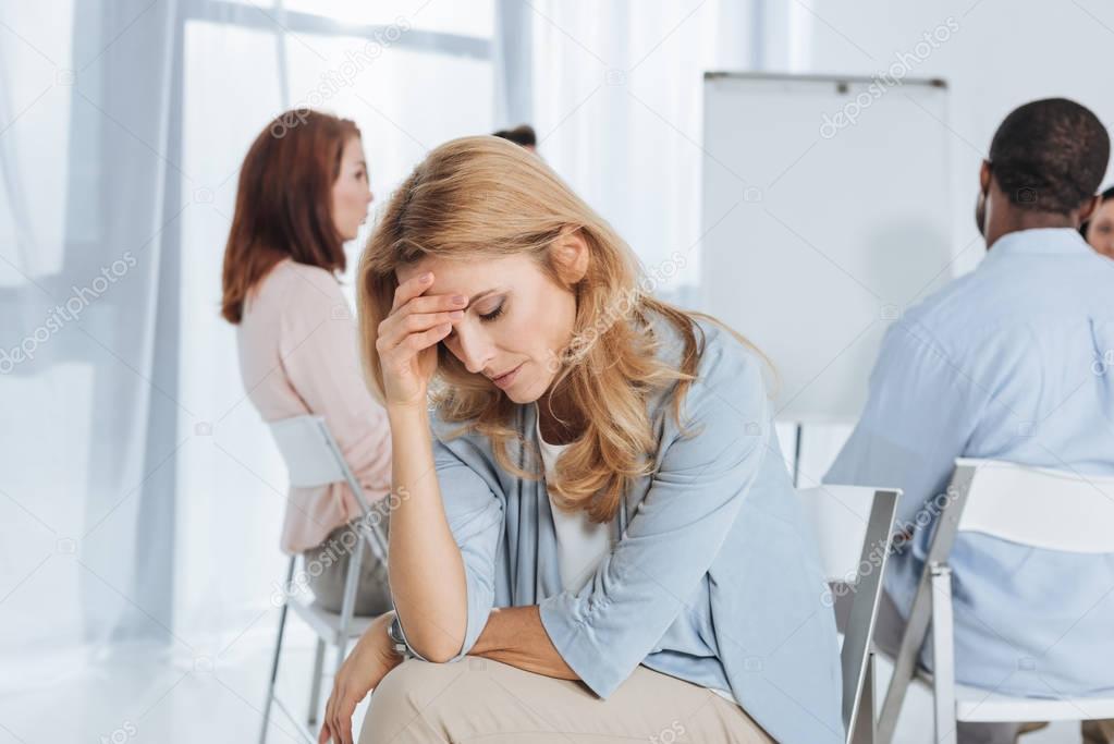 upset mature woman looking down while multiethnic group sitting behind 