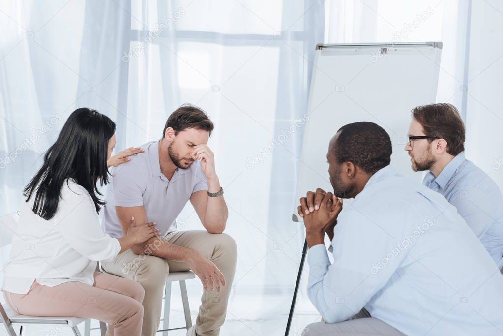 multiethnic middle aged people sitting on chairs and supporting upset man during anonymous group therapy  
