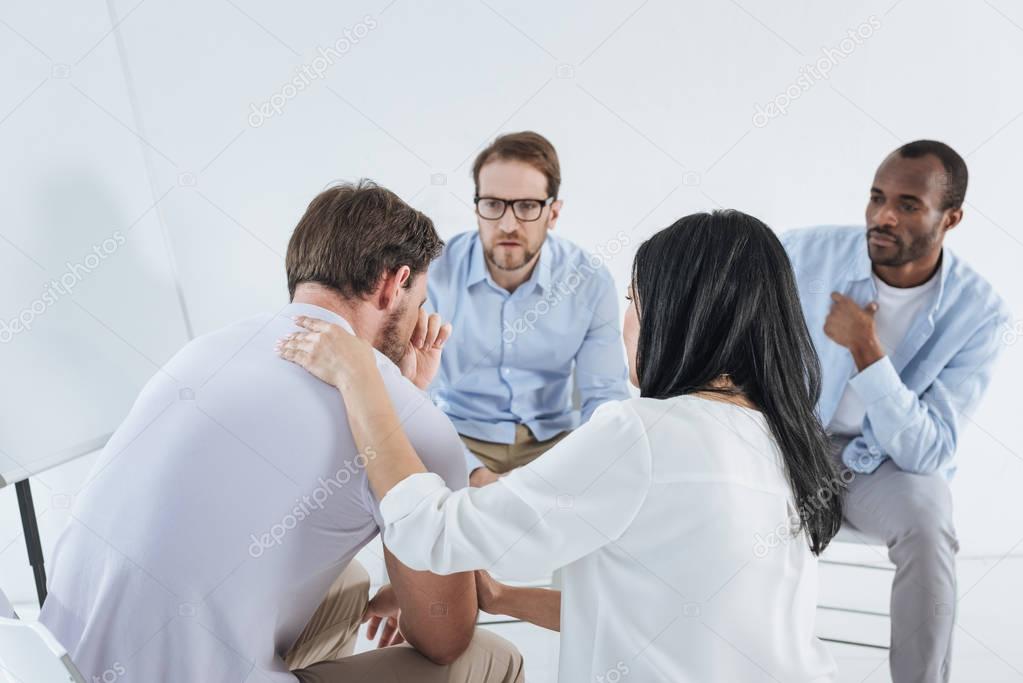 multiethnic mid adult people sitting on chairs and supporting upset man during anonymous group therapy   