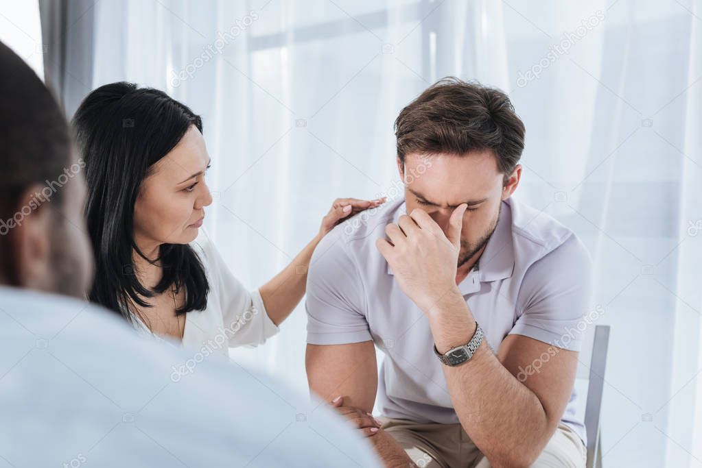 selective focus of mature asian woman supporting crying man during group therapy