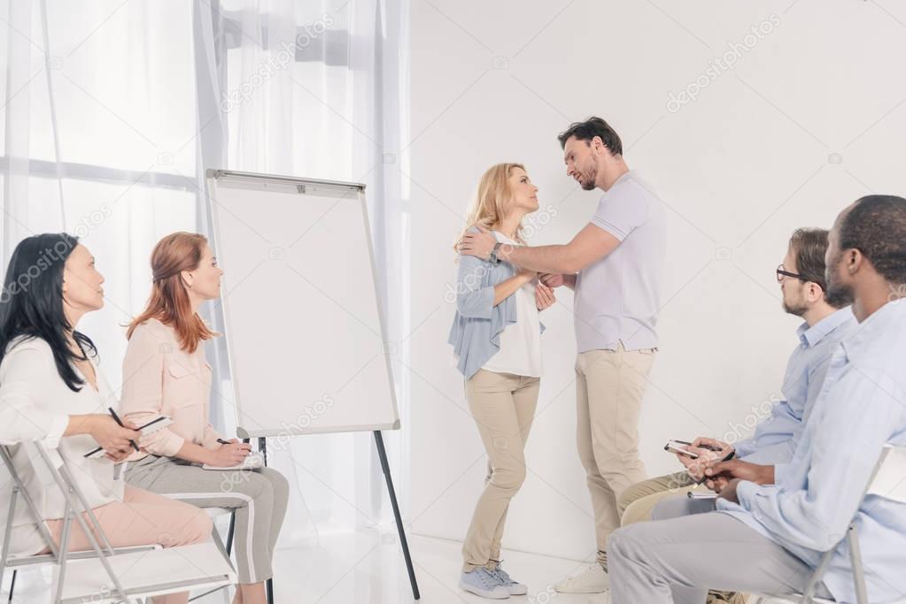 mature couple looking at each other while standing near blank whiteboard and other people sitting on chairs during group therapy 