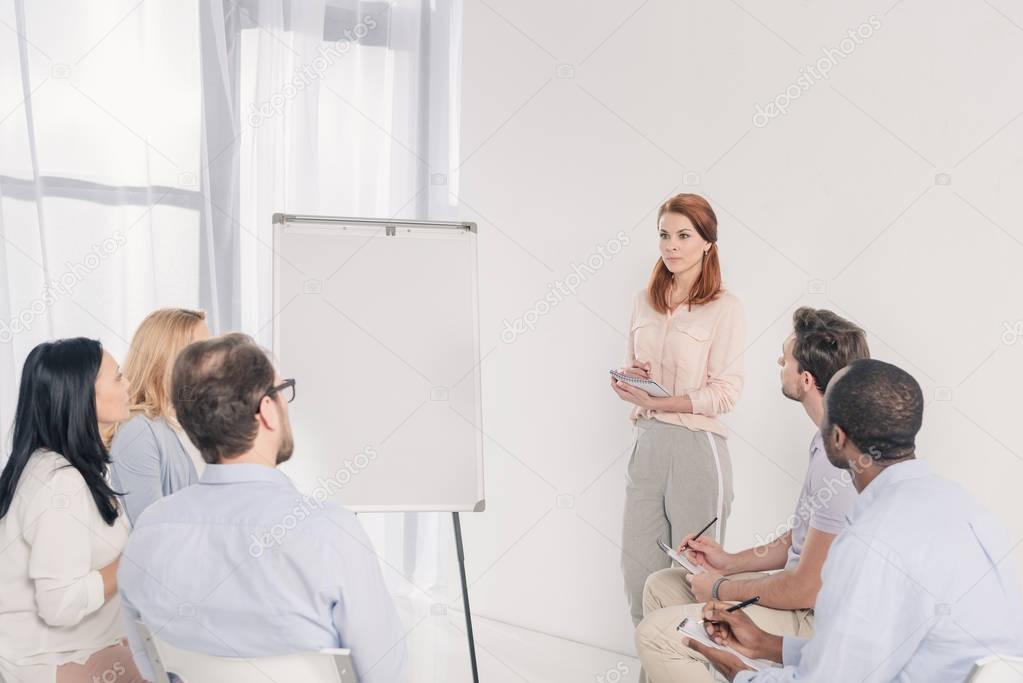 middle aged woman standing near blank whiteboard and looking at multiethnic people during group therapy