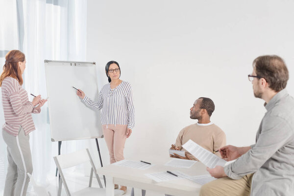multiethnic women with pens standing near blank whiteboard and middle aged men sitting on foreground