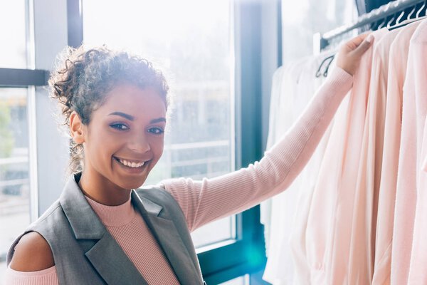 улыбающаяся молодая женщина выбирает новую одежду в магазине
