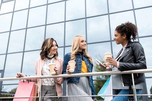 group of young women with shopping bags and coffee to go talking outdoors near mall