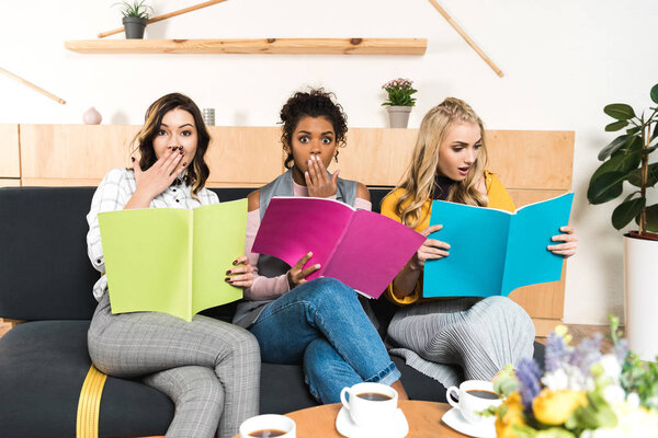shocked young women with magazines sitting on couch in cafe