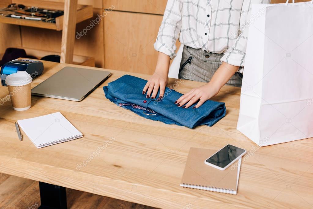 cropped shot of clothing store manager folding shirt to pack it into shopping bag
