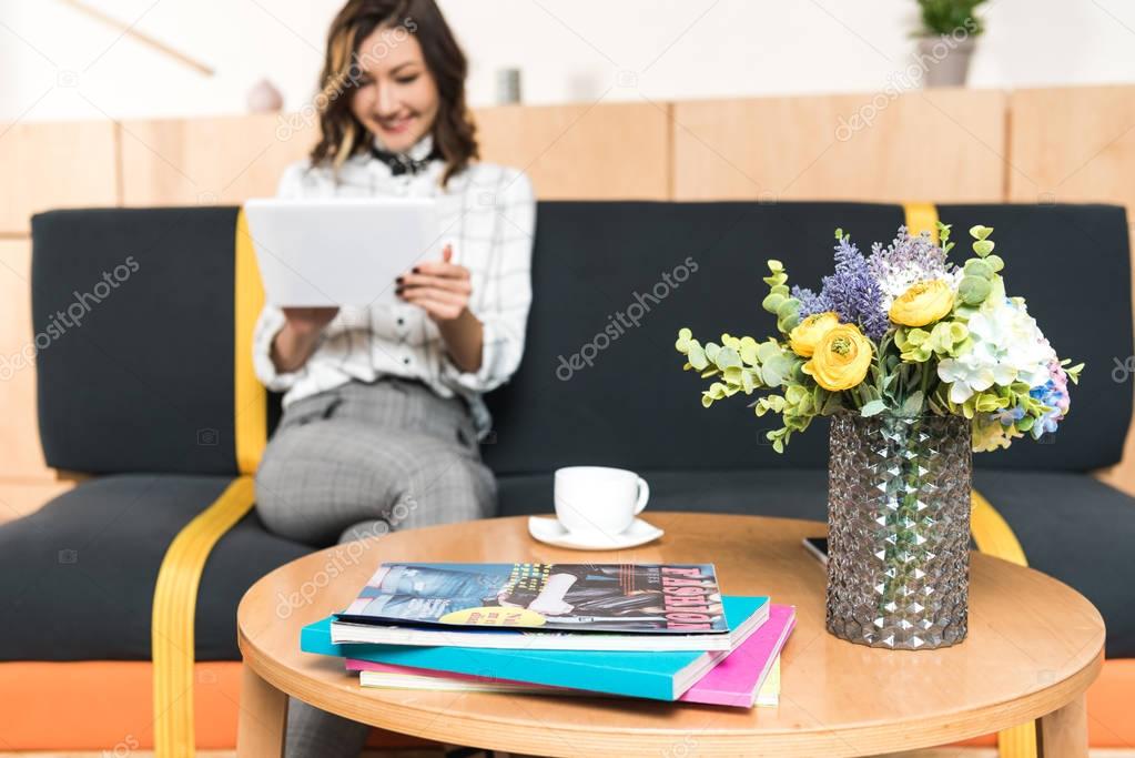 happy young woman using tablet while sitting on couch in cafe with flowers and magazines on table