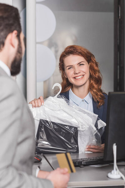 dry cleaning manageress standing at workplace with bag of clothes while customer holding gold credit card