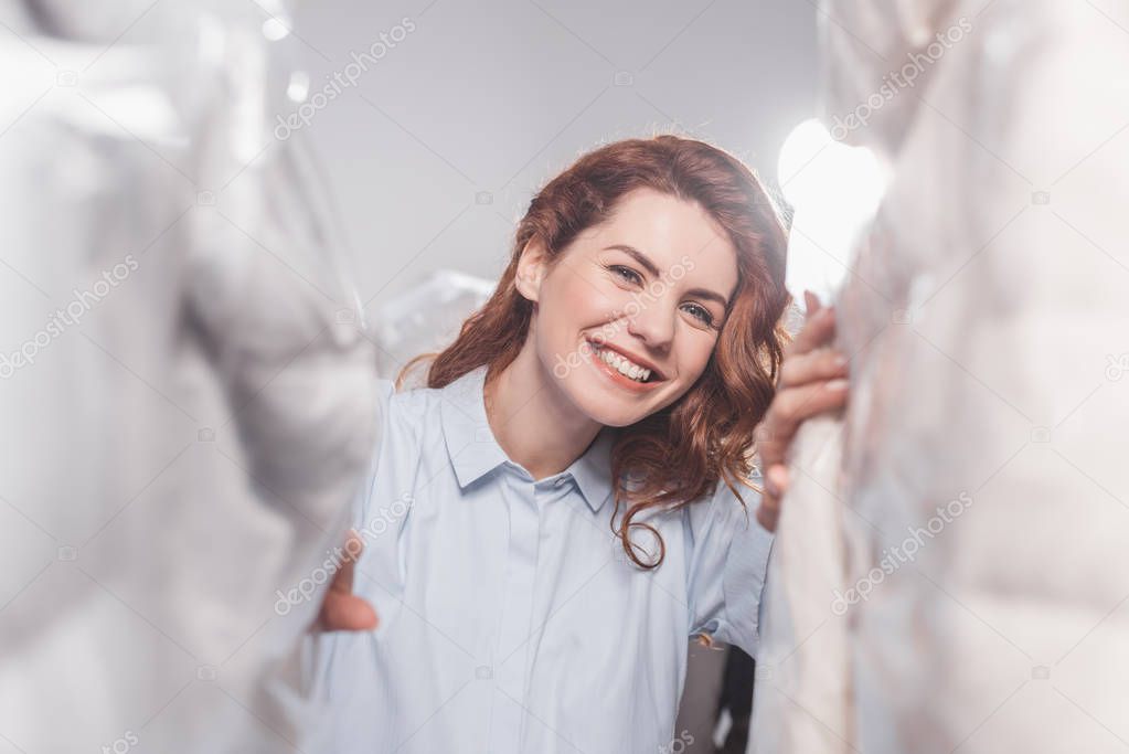 smiling female dry cleaning worker  looking at camera between clothing in plastic bags hanging at warehouse