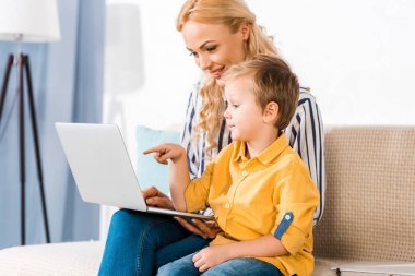 Anne ve küçük oğlu dizüstü birlikte evde kanepede kullanarak gülümseyen