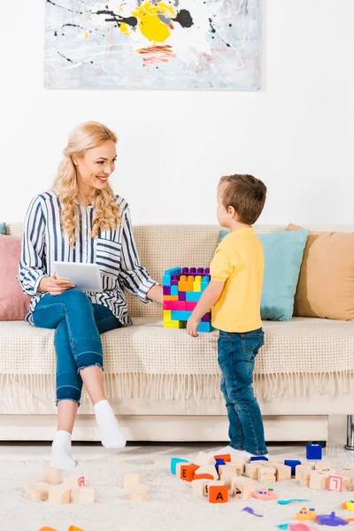 Маленький Мальчик Показывает Игрушки Улыбающейся Матери Табличкой Диване Дома — Бесплатное стоковое фото