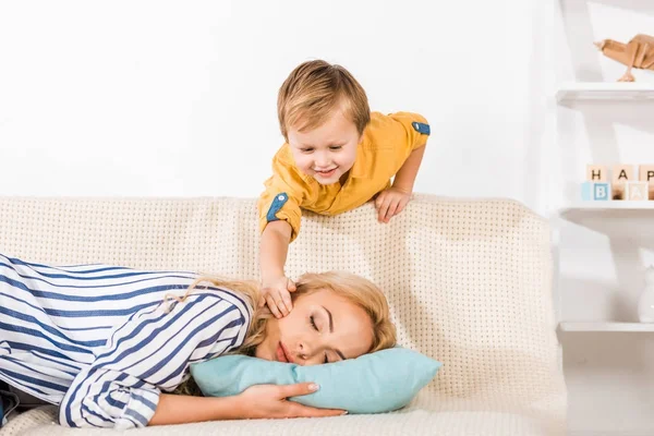 Uśmiechający Się Mały Chłopiec Dotykając Matka Śpi Kanapie Domu — Zdjęcie stockowe