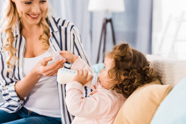 cropped shot of smiling mother feeding adorable infant daughter from bottle at home