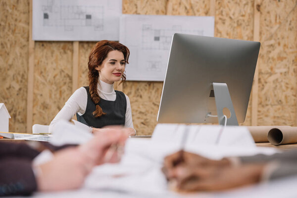 young female architect working with computer at office while colleagues drawong plans on foreground