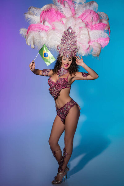 Happy young woman in carnival costume with pink feathers holding flag of Brasil on blue background
