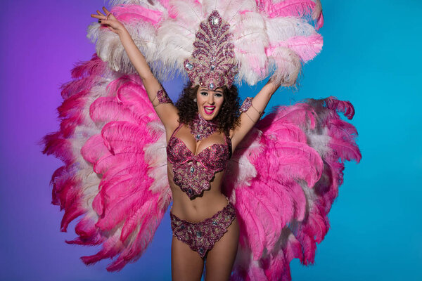 Cheerful woman in carnival costume with pink feathers raising hands isolated on blue background