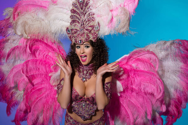 Surprised young woman in carnival costume with pink feathers isolated on blue background
