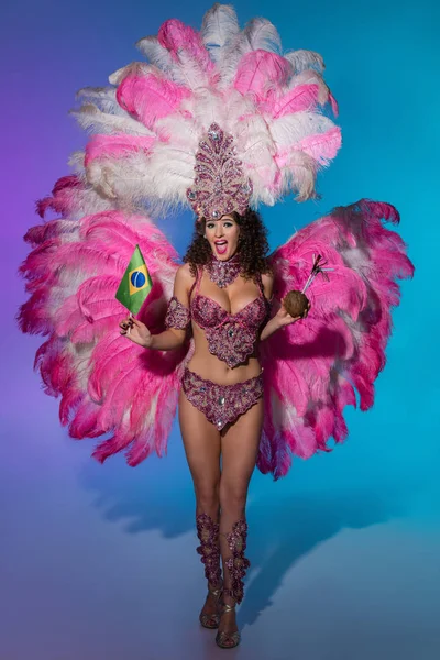 Cheerful woman in carnival costume with pink feathers holding coconut and Brasil flag on blue background