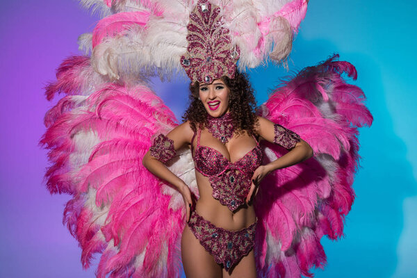 Счастливая девушка в карнавальном костюме с розовыми перьями танцует эмоционально на синем фоне
