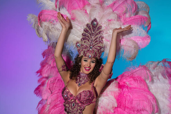 Happy young woman in carnival costume with pink feathers smiling elated isolated on blue background