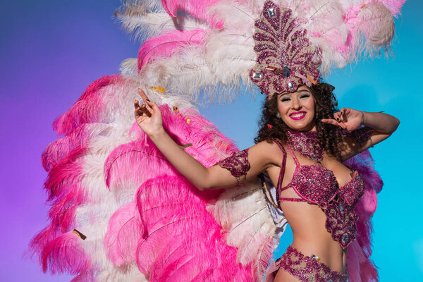 Cheerful woman in carnival costume with pink feathers dancing and smiling isolated on blue background