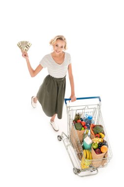 high angle view of young woman holding dollar banknotes and smiling at camera while standing with shopping trolley full of grocery isolated on white clipart