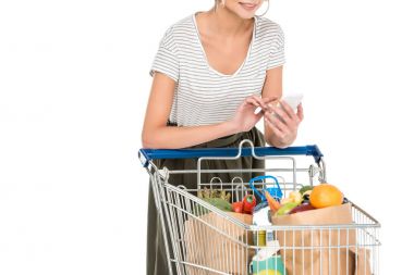 cropped shot of young woman using smartphone while leaning at shopping trolley with grocery bags isolated on white