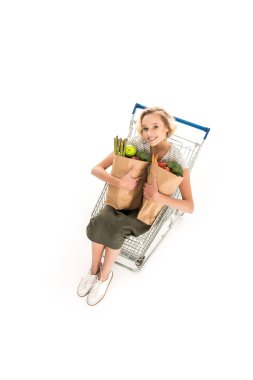 high angle view of girl smiling at camera while holding grocery bags and sitting in shopping trolley isolated on white clipart