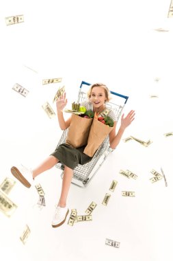 excited young woman with grocery bags sitting in shopping trolley and throwing dollar banknotes isolated on white clipart