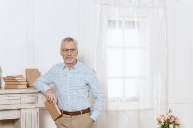 Senior man stands by fireplace in room holding a book clipart
