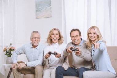 Middle aged men and women sitting on sofa and playing game console using joysticks clipart
