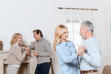 Middle aged men and women dancing and talking by fireplace in cozy room clipart