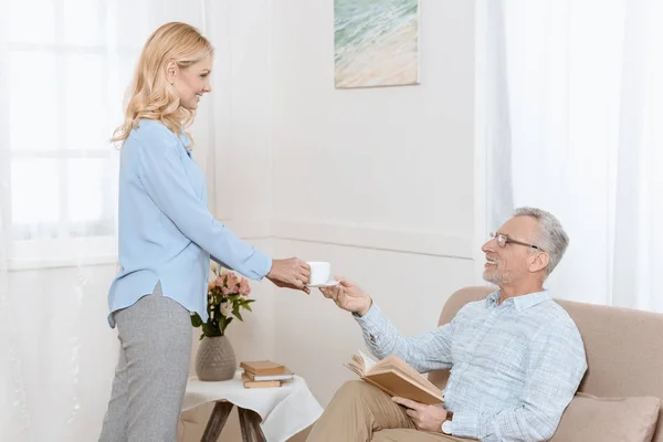 Älterer Mann Liest Buch Während Frau Ihm Tee Hellen Raum — kostenloses Stockfoto