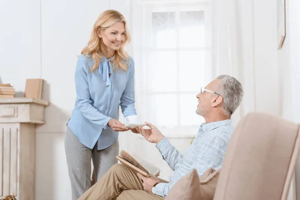 Mature Man Reading Book While Woman Serves Him Tea Light — Stock Photo, Image