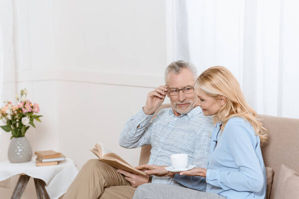 Middle aged woman and man reading book together on sofa in light room