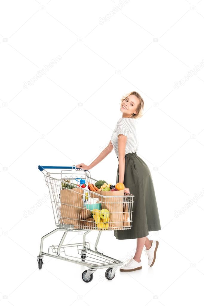 cheerful young woman standing with shopping trolley and smiling at camera isolated on white   