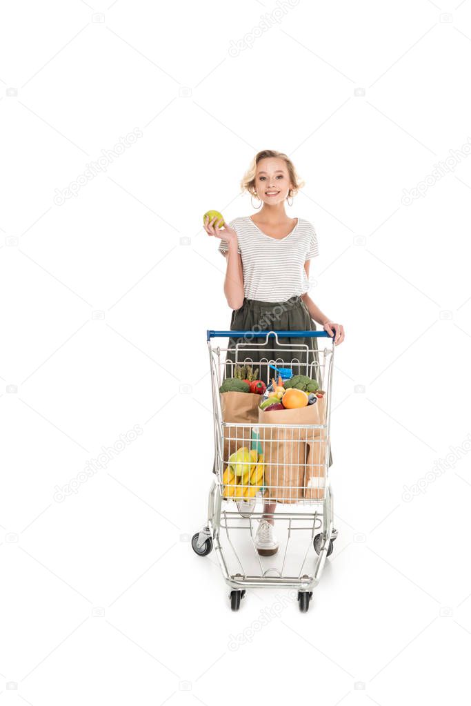 beautiful young woman holding apple and smiling at camera while standing with shopping trolley with grocery bags isolated on white 