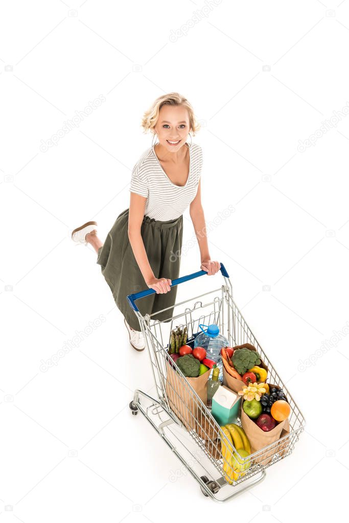 high angle view of cheerful young woman smiling at camera while pushing shopping trolley with grocery bags isolated on white