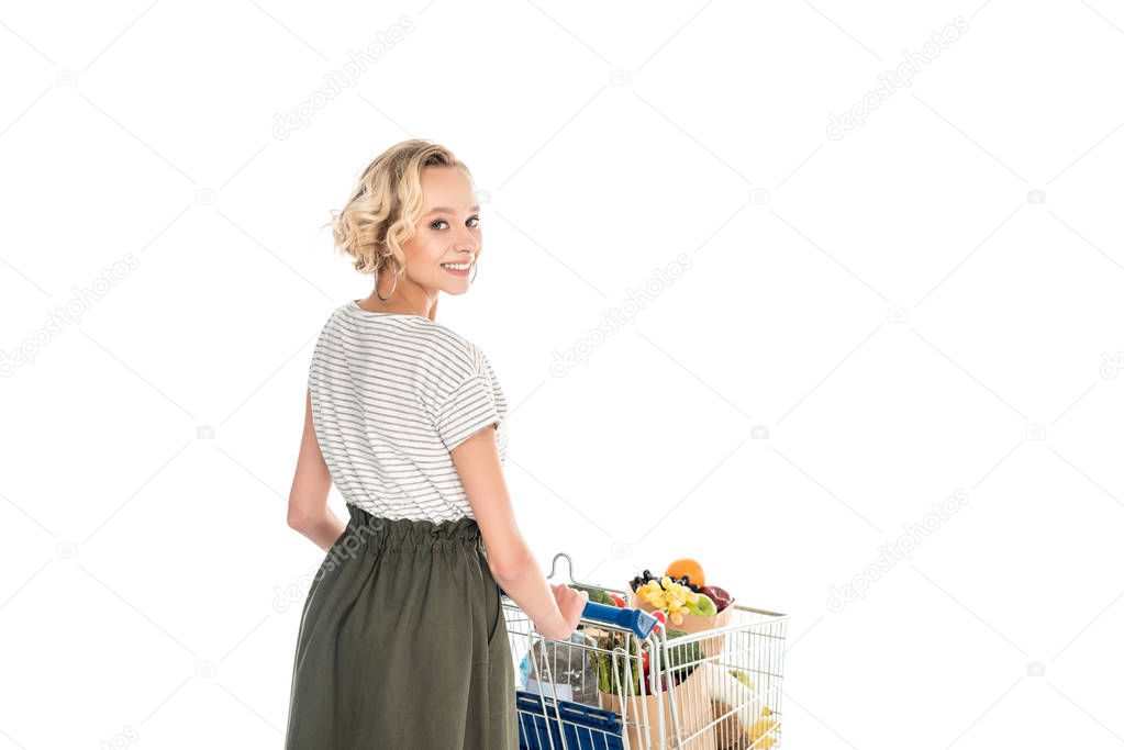 beautiful girl smiling at camera while pushing shopping trolley with grocery bags and plastic bottle of water isolated on white