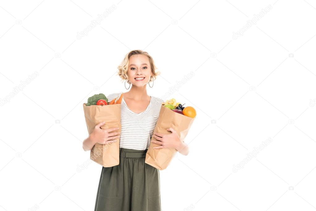 young woman holding paper bags with fruits and vegetables and smiling at camera isolated on white